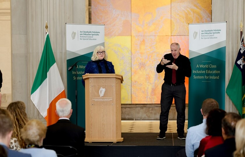 A speaker at a podium with an Irish flag and a sign language interpreter on stage at an NCSE conference