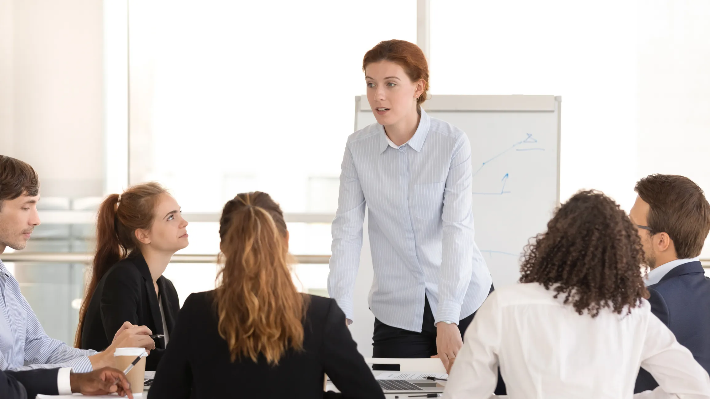 Person standing and speaking with colleagues seated around a table