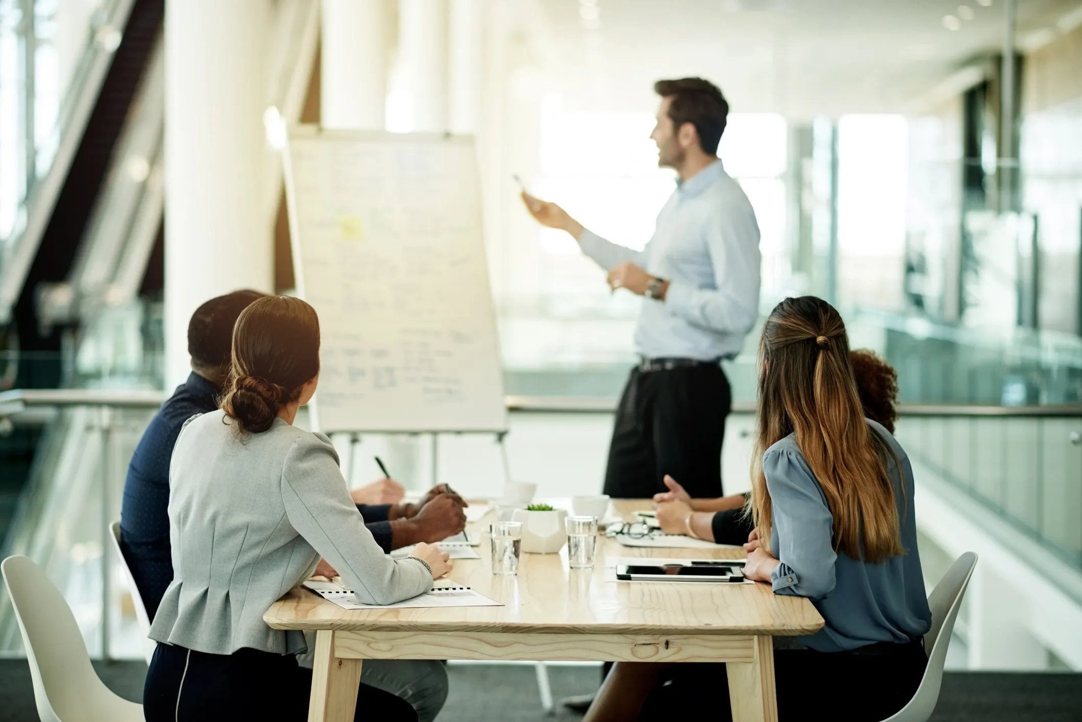 Meeting with a presenter pointing at a flip chart while colleagues listen and take notes