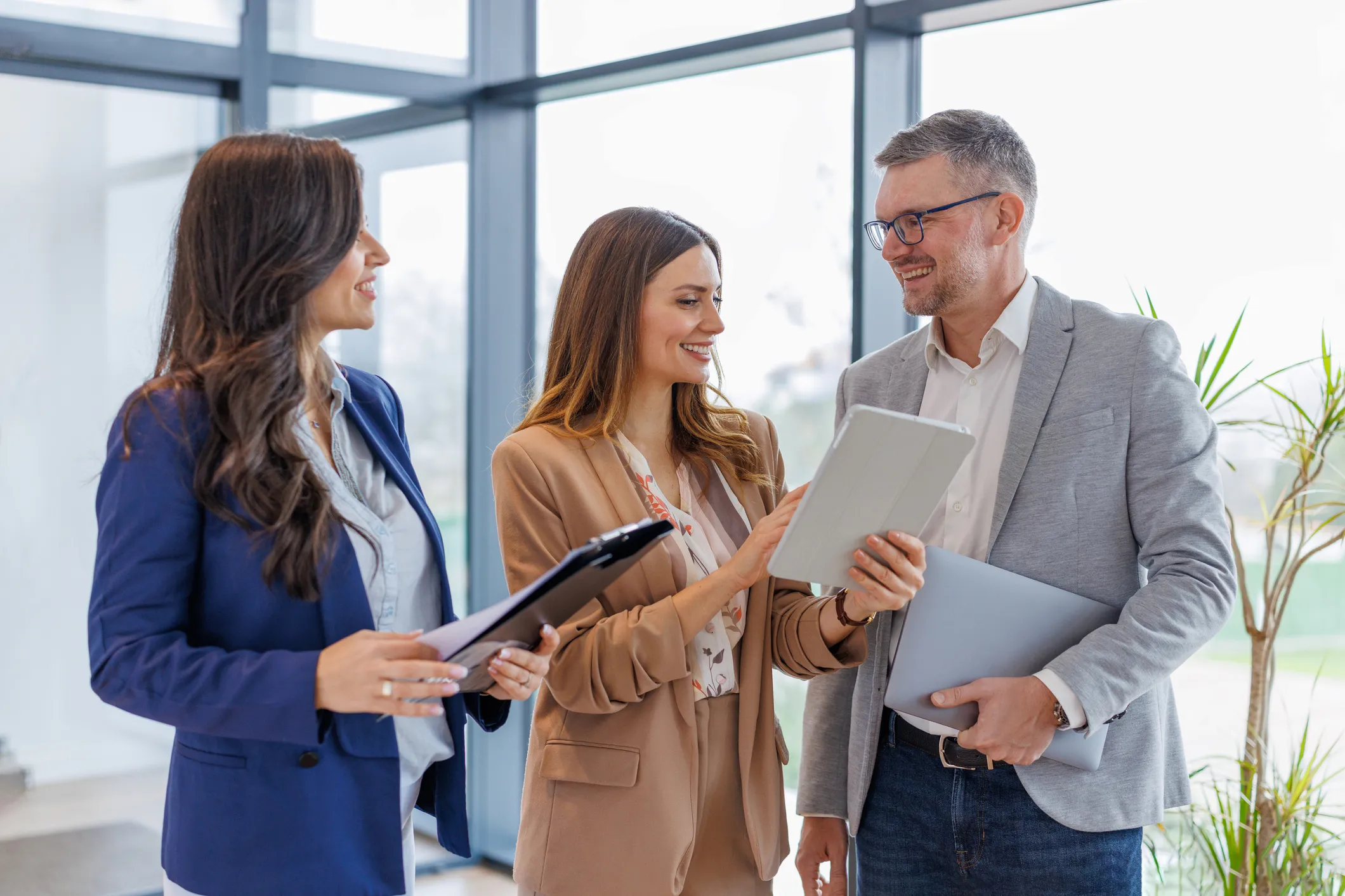 Three people standing having a discussion while holding a tablet and documents in an office