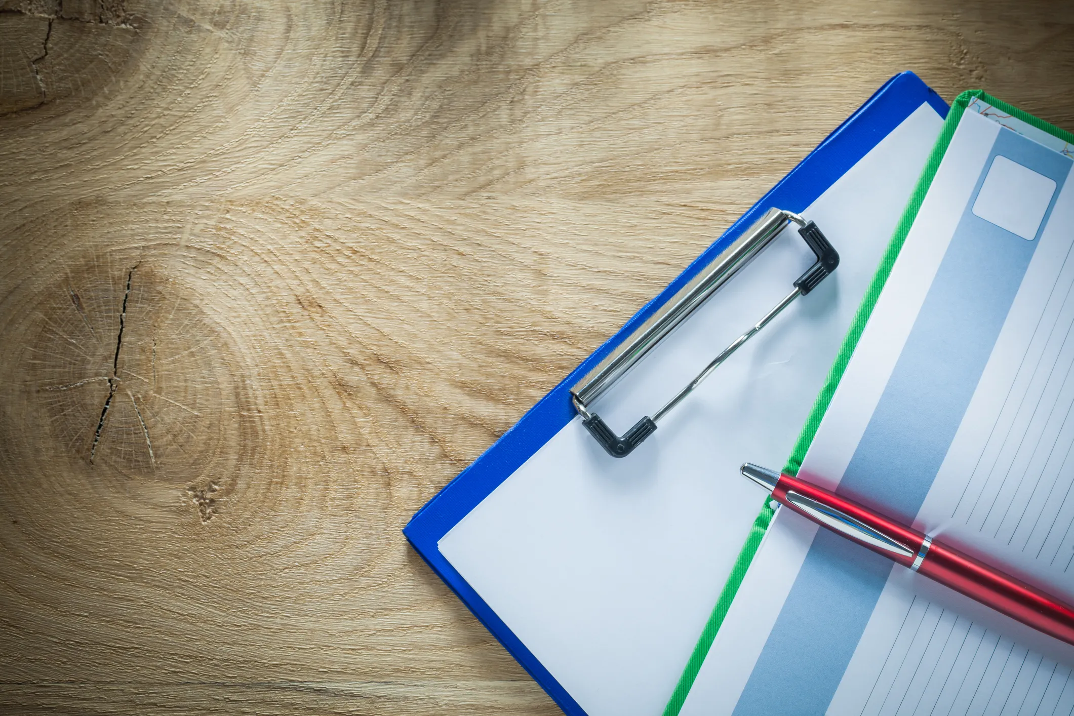 A clipboard holding documents sits on a wooden surface, with a red pen resting on top of the papers.
