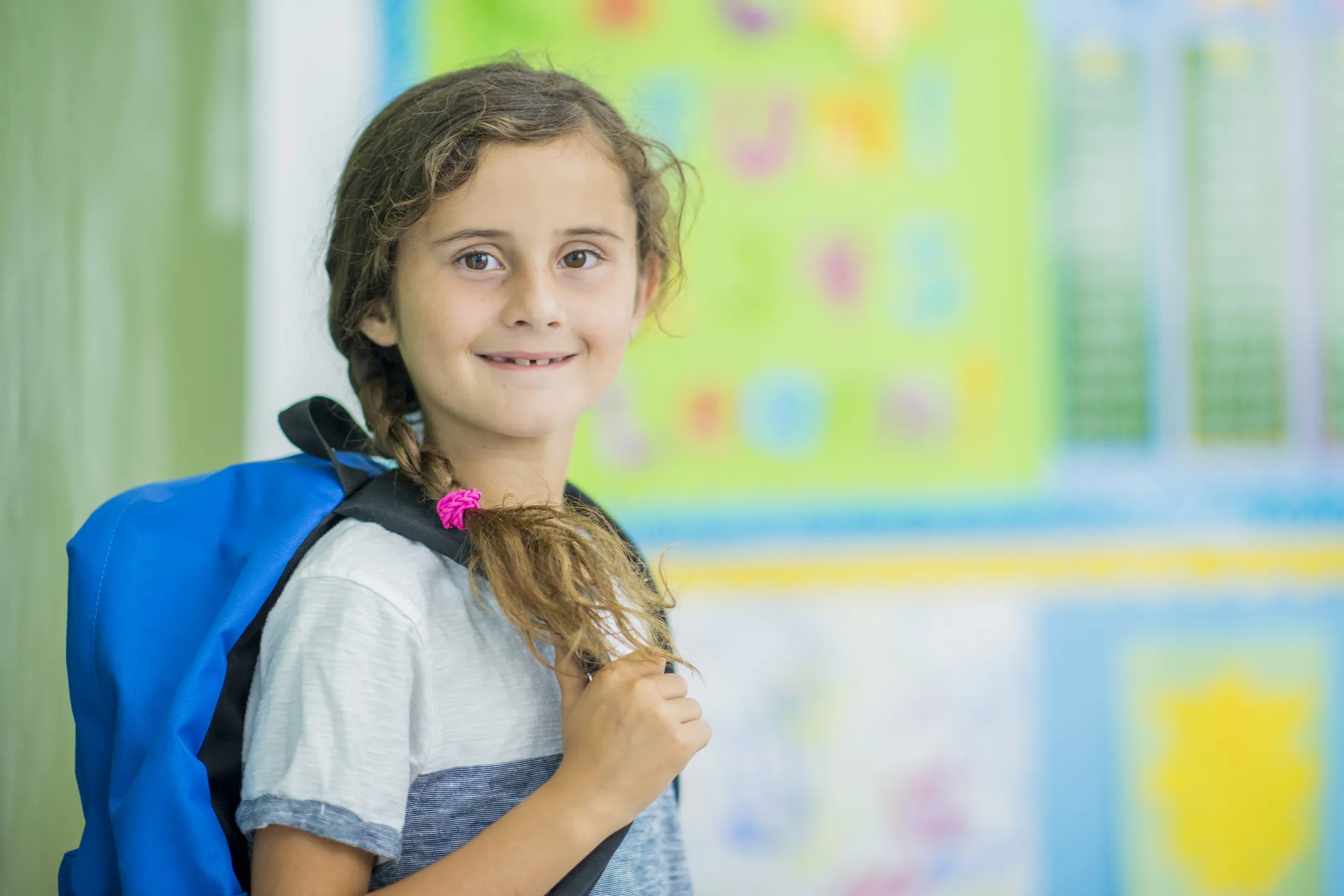 A child preparing to leave a classroom wearing a backpack, holding one of the straps, with colourful classroom-style posters blurred in the background.