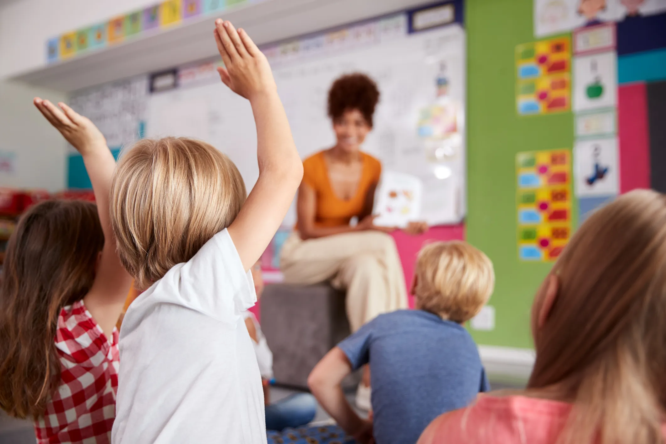 Children sitting on the floor in a classroom, raising their hands while a teacher reads a book at the front