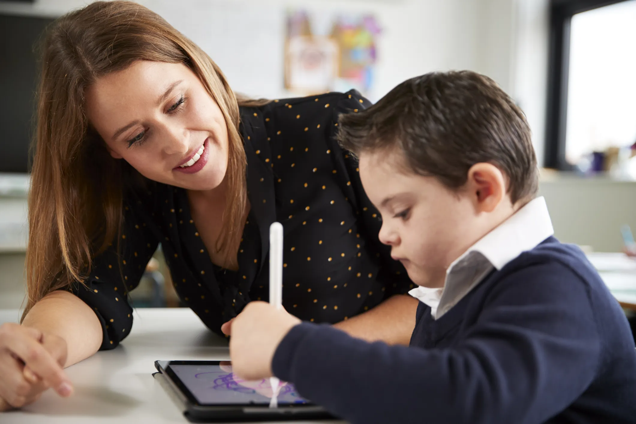 Teacher assisting a young student with a tablet in a classroom
