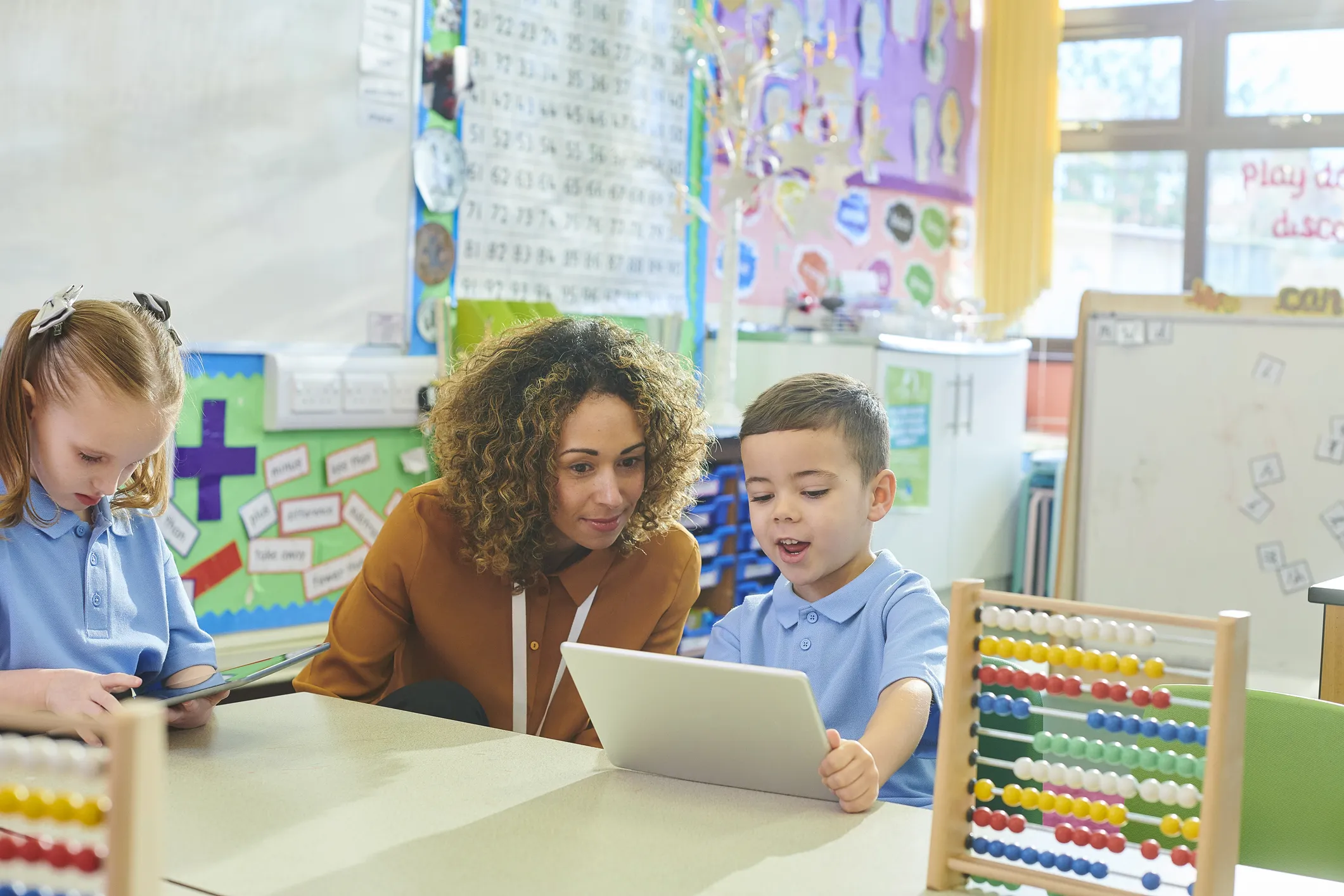 Teacher assisting two young students with tablets in a classroom