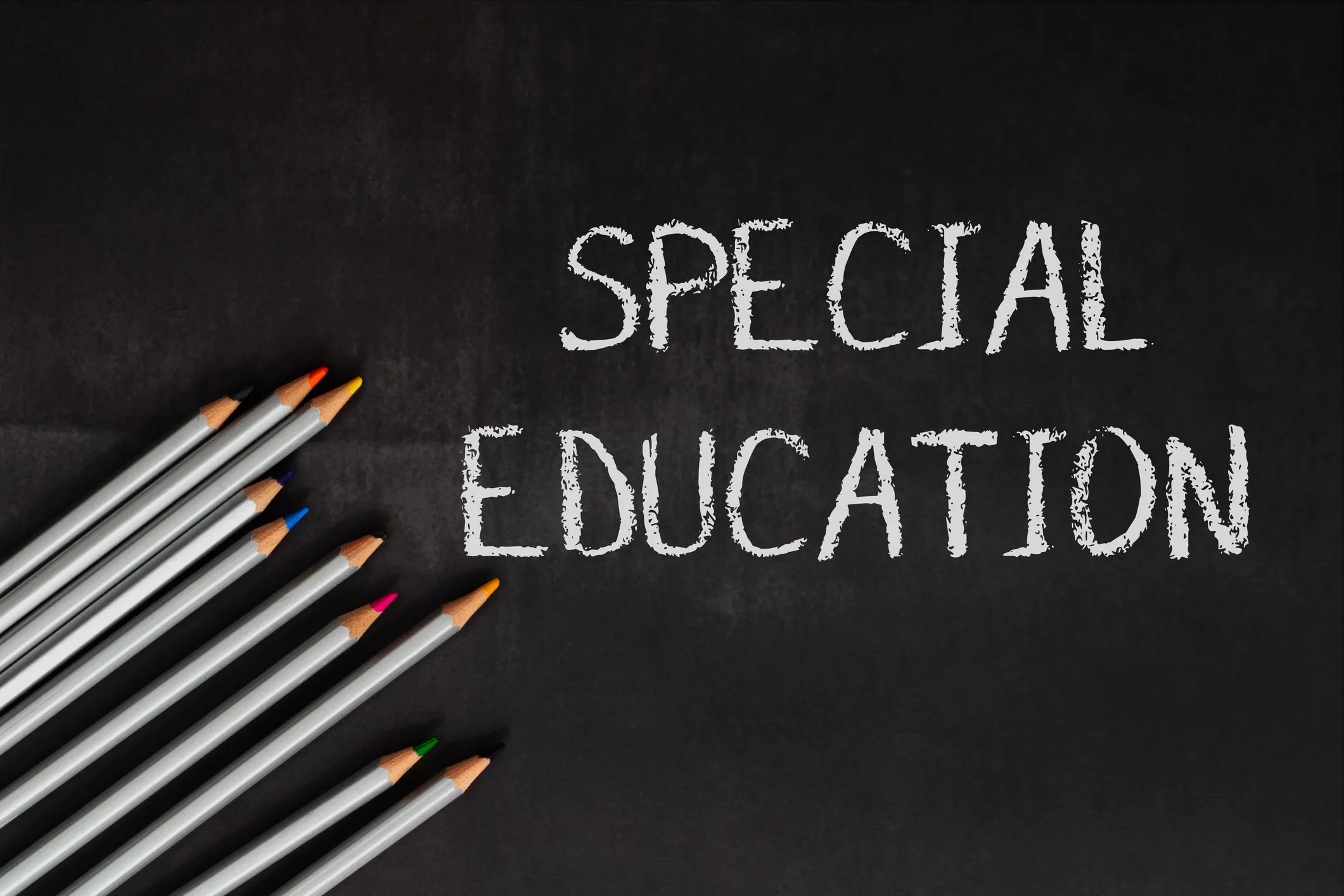 The words “SPECIAL EDUCATION” written in white chalk on a blackboard, with a row of coloured pencils arranged along the left side.