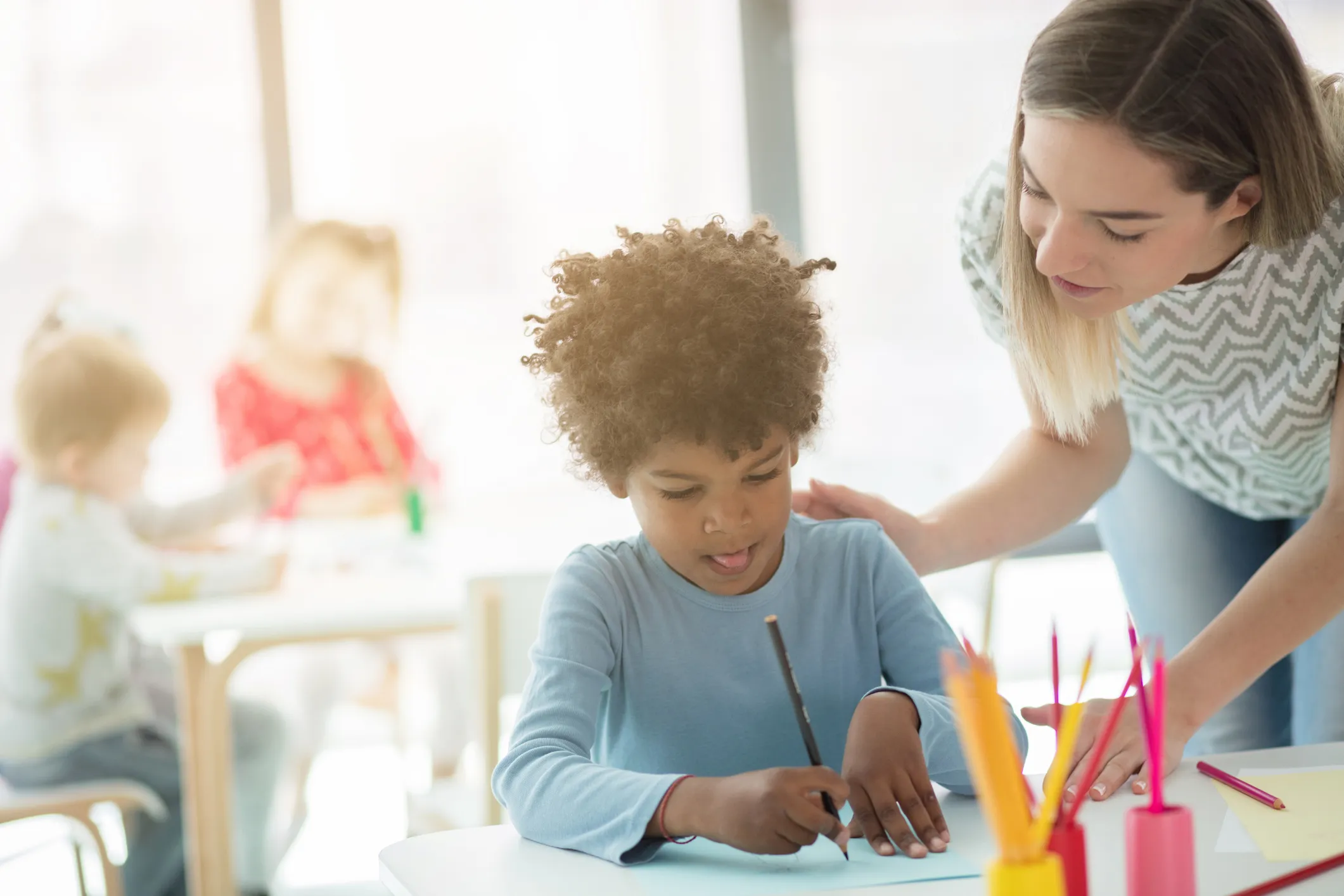 Child writing at a desk with teacher assisting in a classroom