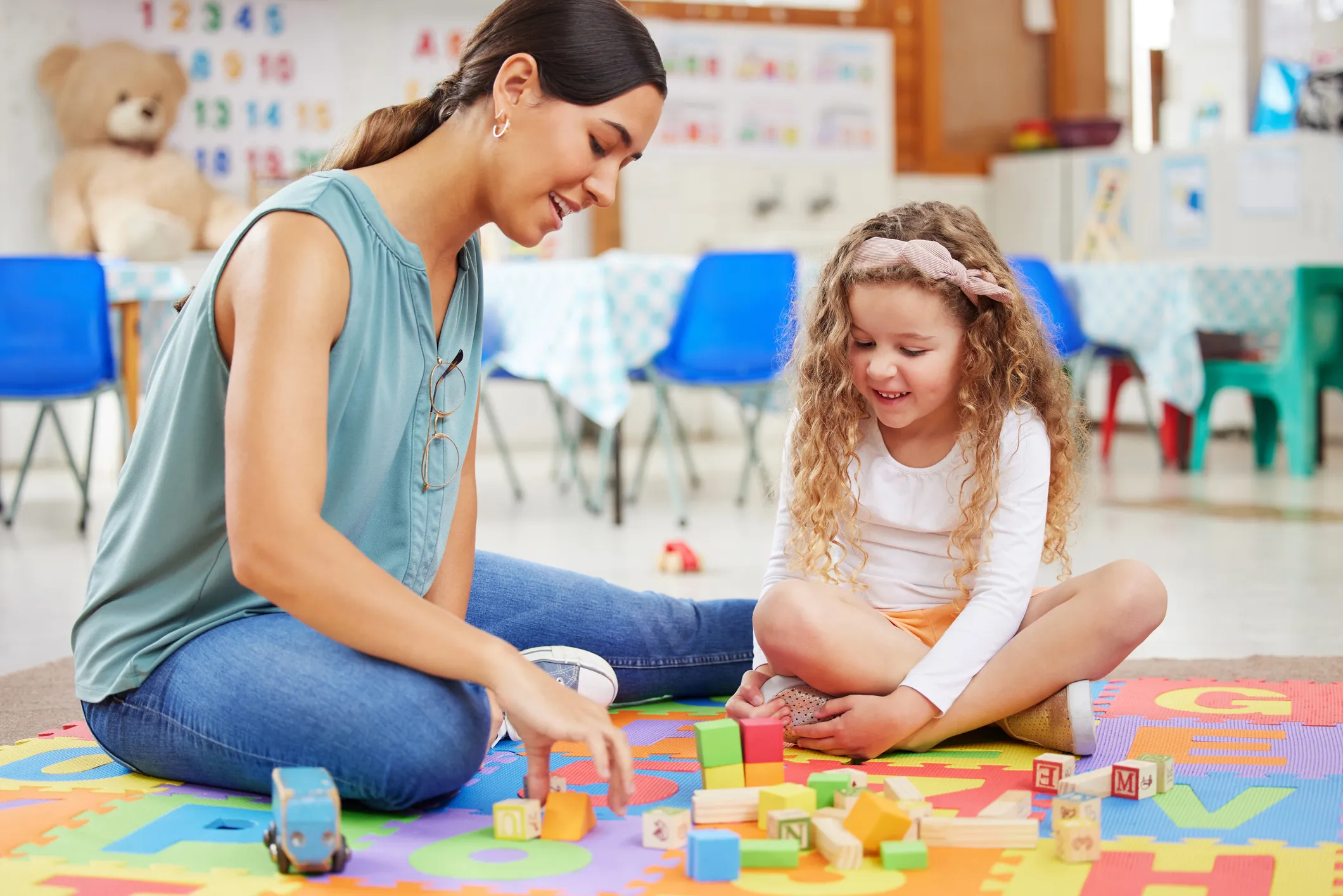 An adult and a young child are sitting on a colorful foam mat in a classroom setting playing